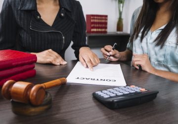 black-woman-pointing-at-document-near-lady-with-pen-at-table-with-calculator-and-gavel
